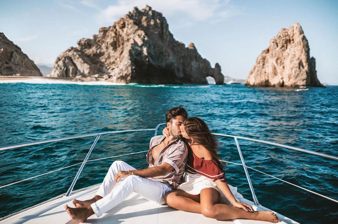 Casal em iate de luxo beijando-se com o icônico Arco de Los Cabos ao fundo, cenário romântico para sua viagem.